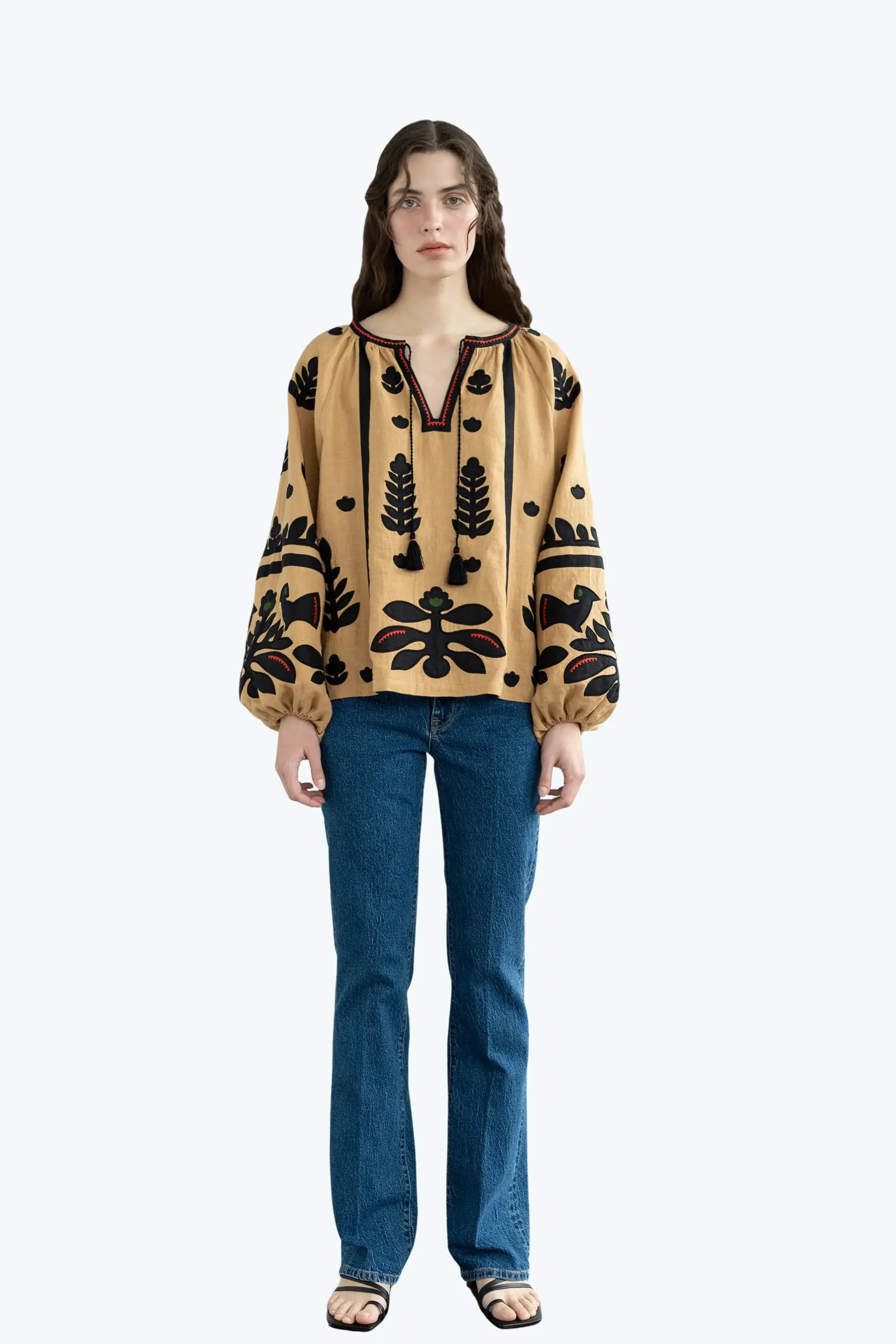 A model stands against a light gray background wearing a beige long-sleeve blouse with black embroidered patterns and blue jeans.