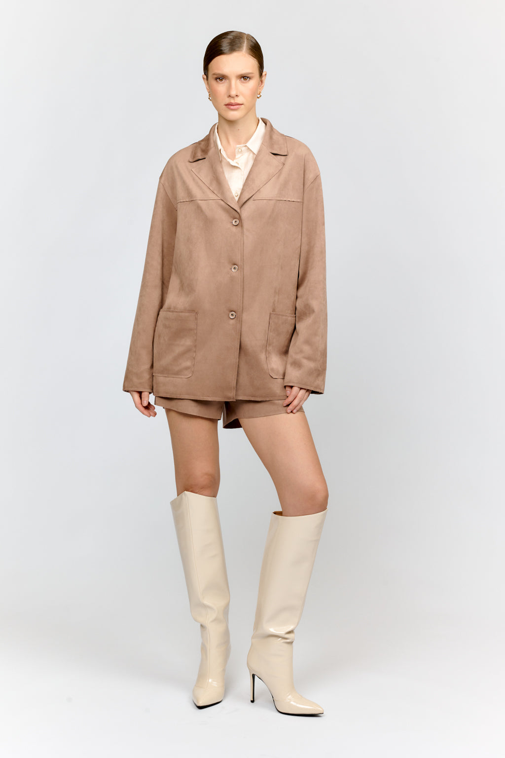 Woman wearing a beige coat, white shirt, shorts, and cream knee-high boots on a light gray background