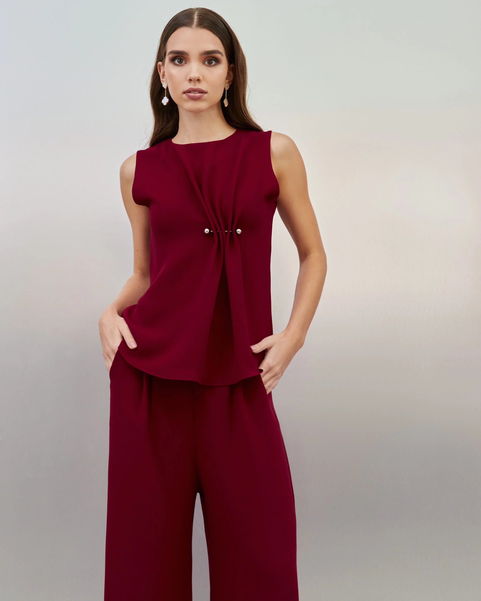 A woman modeling a sleeveless deep burgundy jumpsuit with a gathered front detail and small decorative buttons, set against a neutral gray studio background.