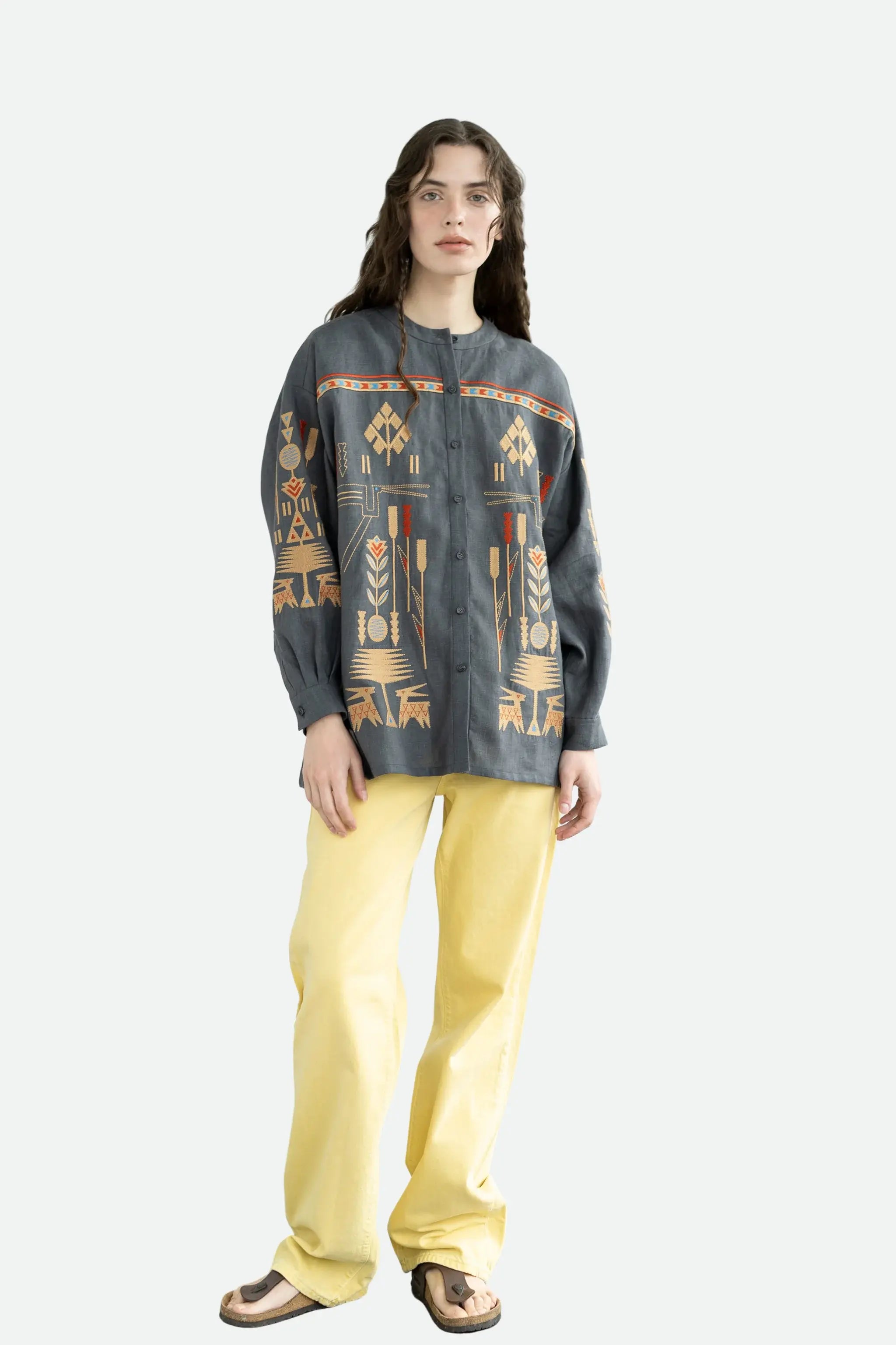 A model stands against a light gray studio background wearing a dark gray button-up shirt with a colorful geometric tribal-style print in orange, beige, and red, paired with bright yellow pants and brown sandals.