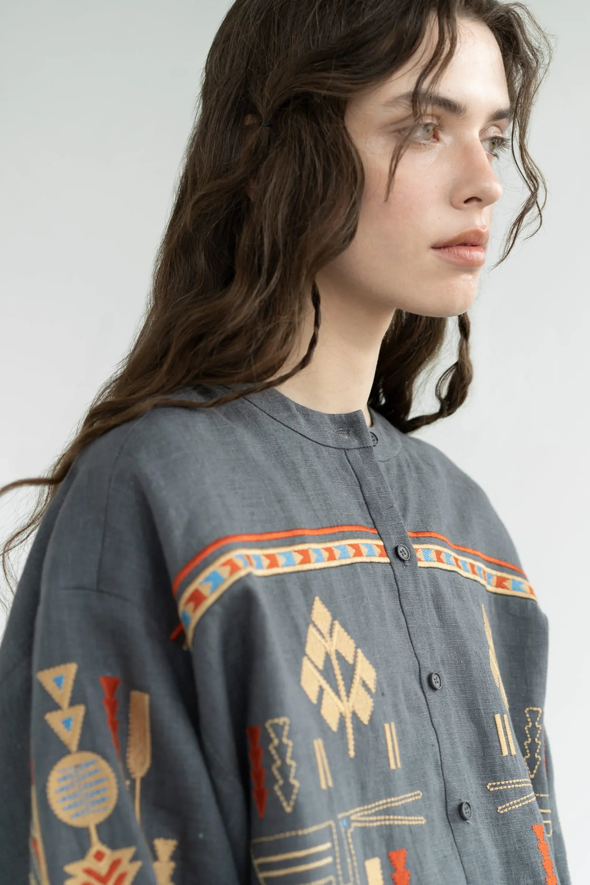 A close-up side profile of a woman wearing a gray button-front shirt with colorful geometric and tribal-inspired patterns in beige, orange, and blue, against a plain light gray background.