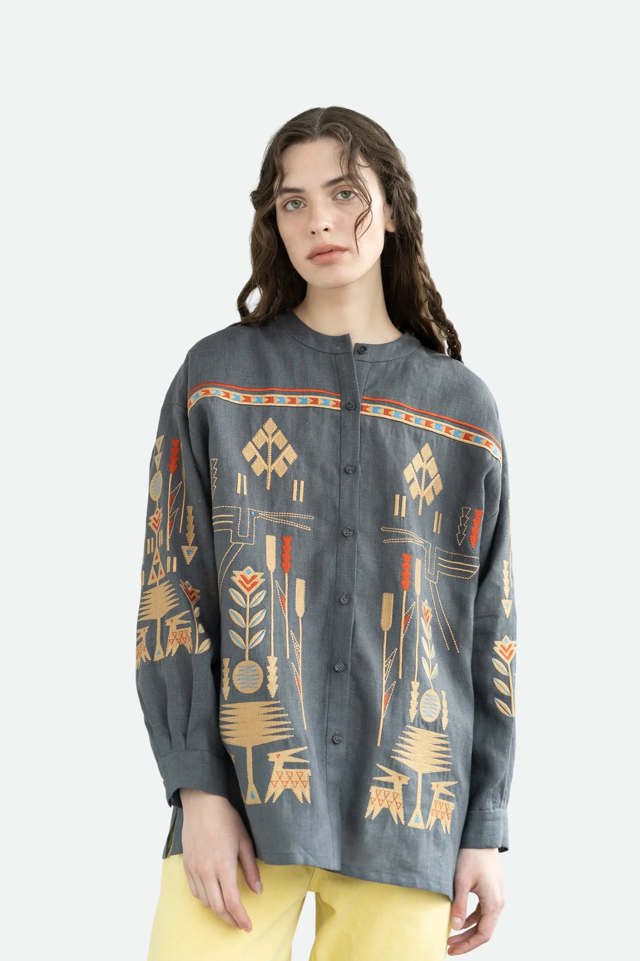 A woman modeling a dark gray button-up shirt with a colorful geometric and tribal-inspired print in orange, beige, and red, paired with yellow pants against a light gray studio background.