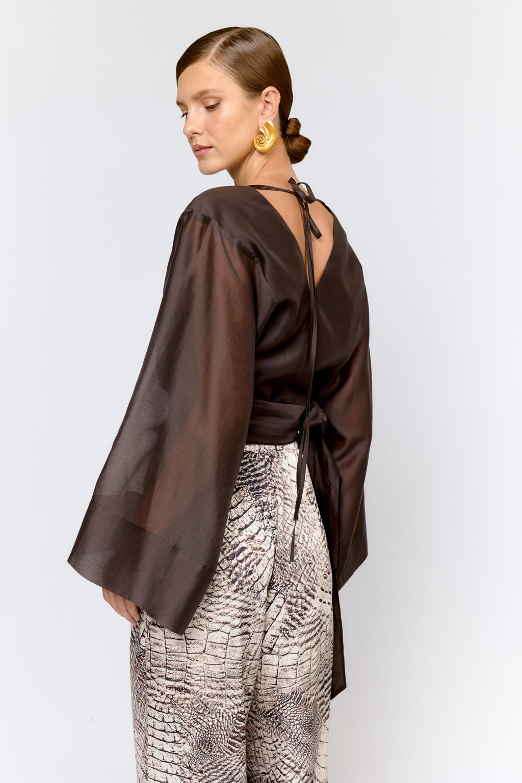 Woman wearing a brown blouse with a back tie and patterned pants on a white background