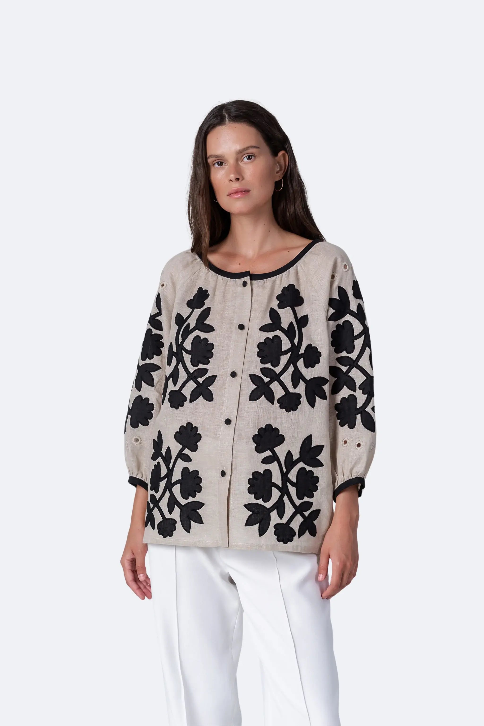 A woman modeling a beige linen button-up cardigan with black floral embroidery, paired with white trousers, against a light gray studio background.