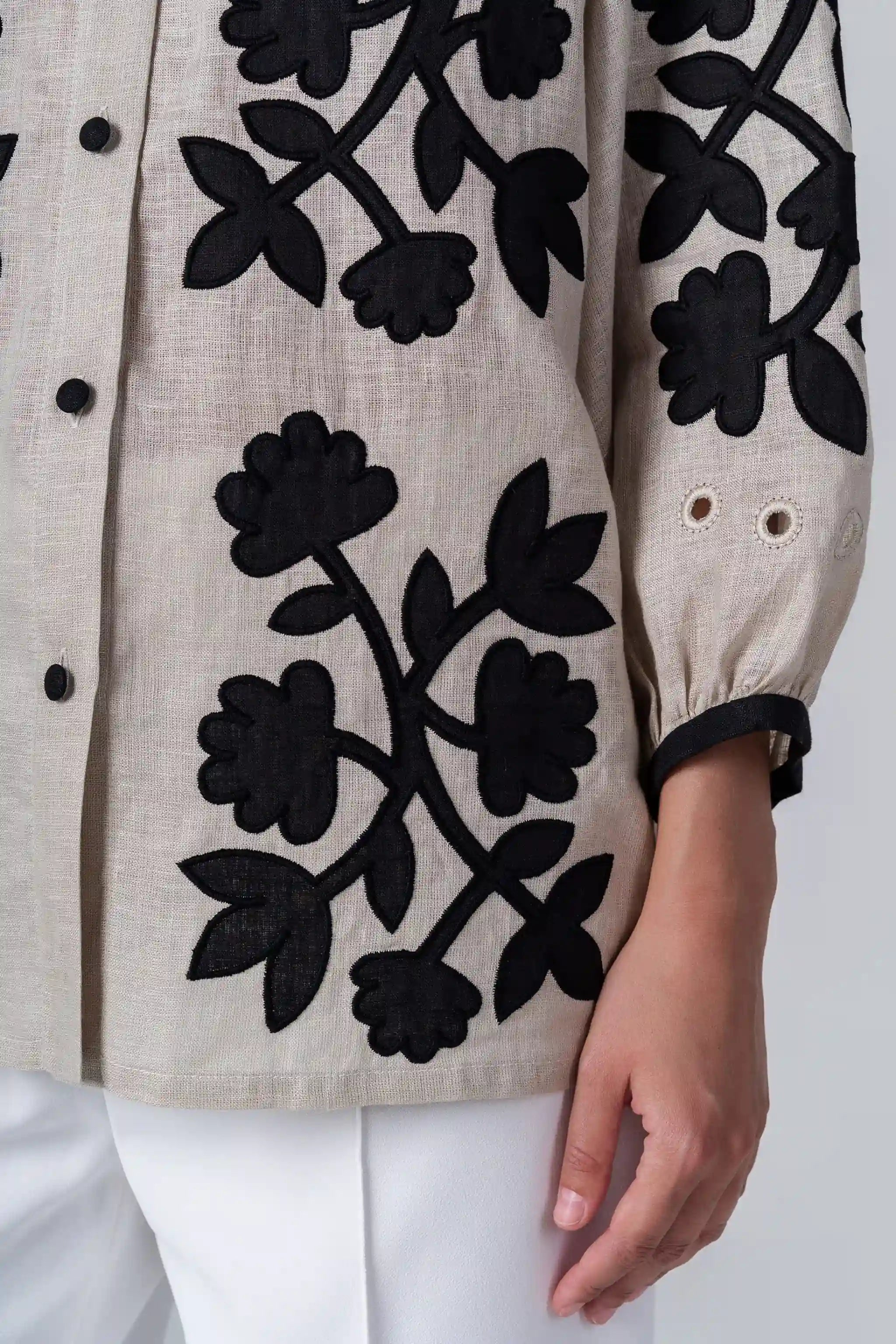 Close-up of a beige linen button-up shirt with black floral embroidery and a model's hand resting near the hem.