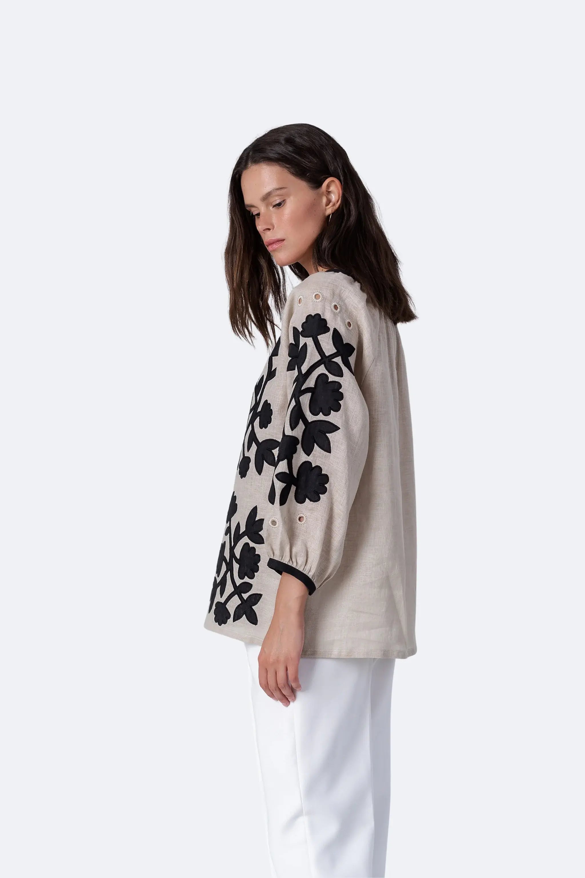 A woman shown in side profile wearing a light beige blouse with prominent black floral embroidery on the sleeves and black trim at the cuffs, paired with white pants, against a plain light gray studio background.