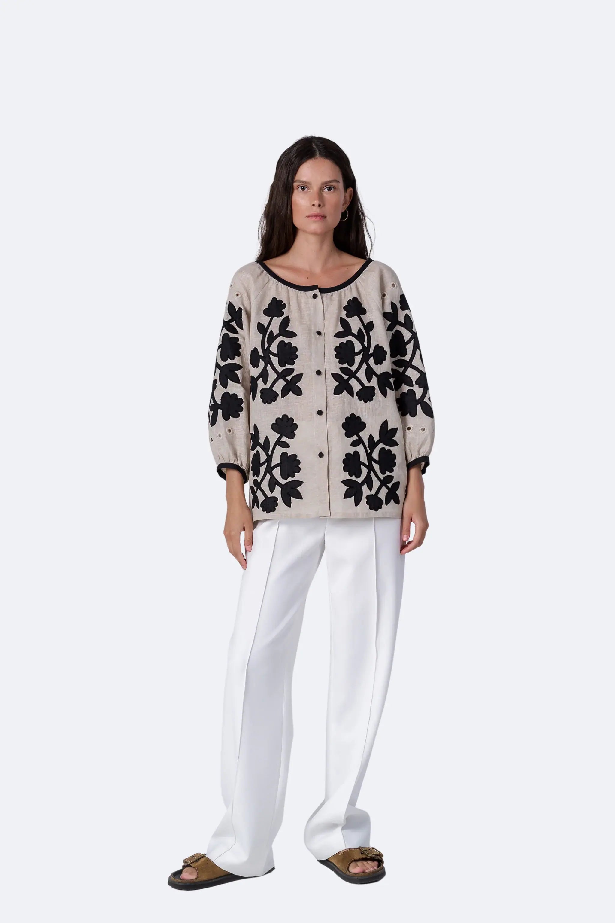 A model stands against a light gray studio background wearing a beige knit cardigan with black floral embroidery, white wide-leg pants, and brown slide sandals.