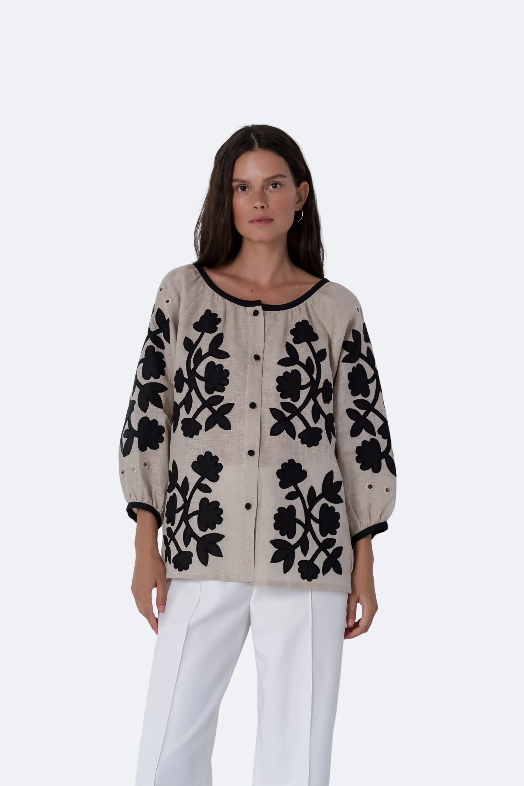 A woman modeling a beige button-front cardigan with black floral embroidery and black trim, paired with white pants, against a light gray studio background.