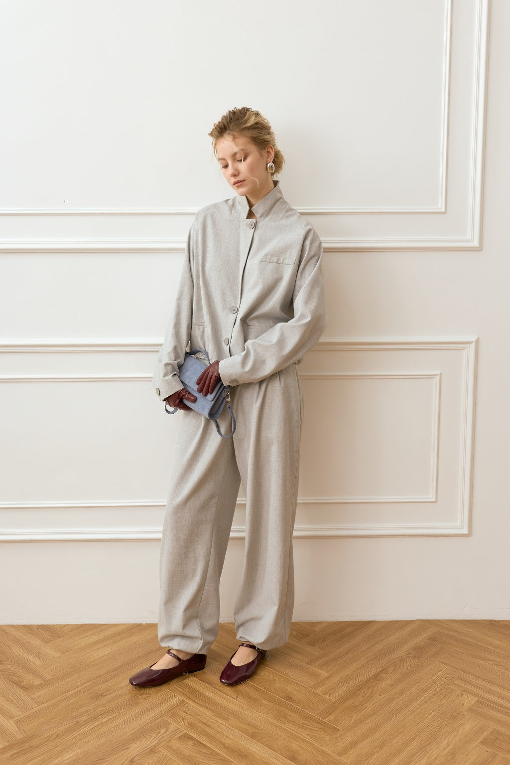 Woman in a light gray outfit standing against a white paneled wall.