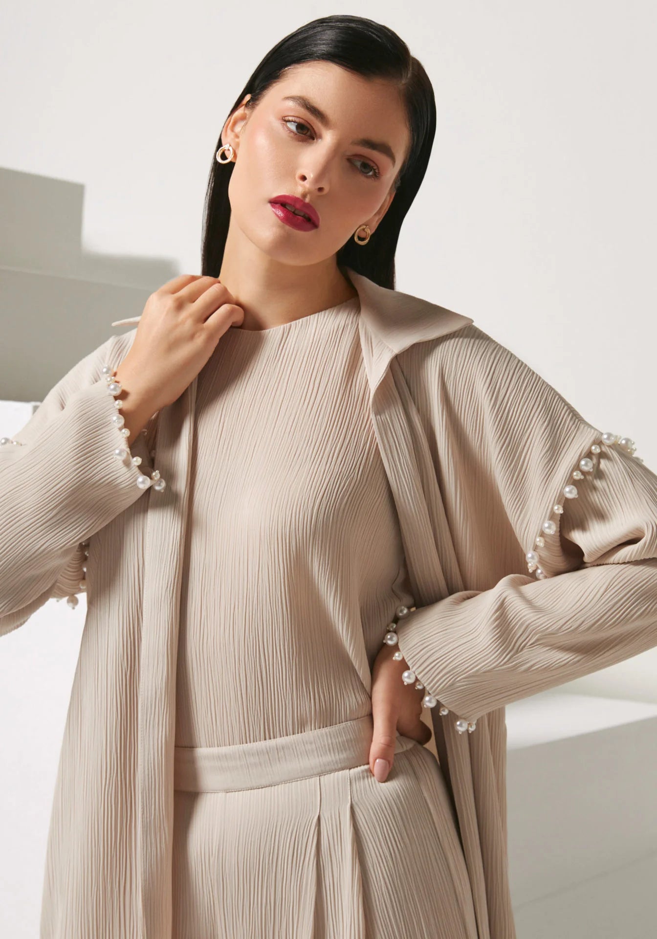 A woman modeling a beige pleated long-sleeve dress with a matching belt and a draped pleated cardigan featuring pearl trim at the cuffs, set against a minimalist white background.