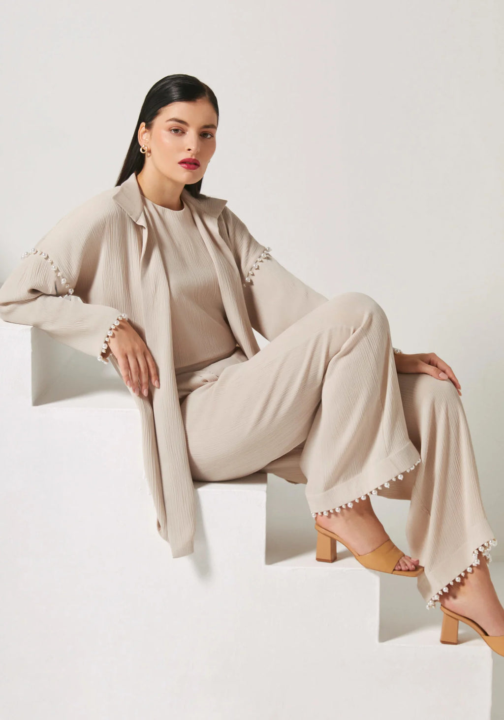 A woman models a beige two-piece outfit consisting of a long-sleeve button-front jacket with scalloped edges and matching wide-leg trousers, styled with tan heeled sandals on white geometric steps.