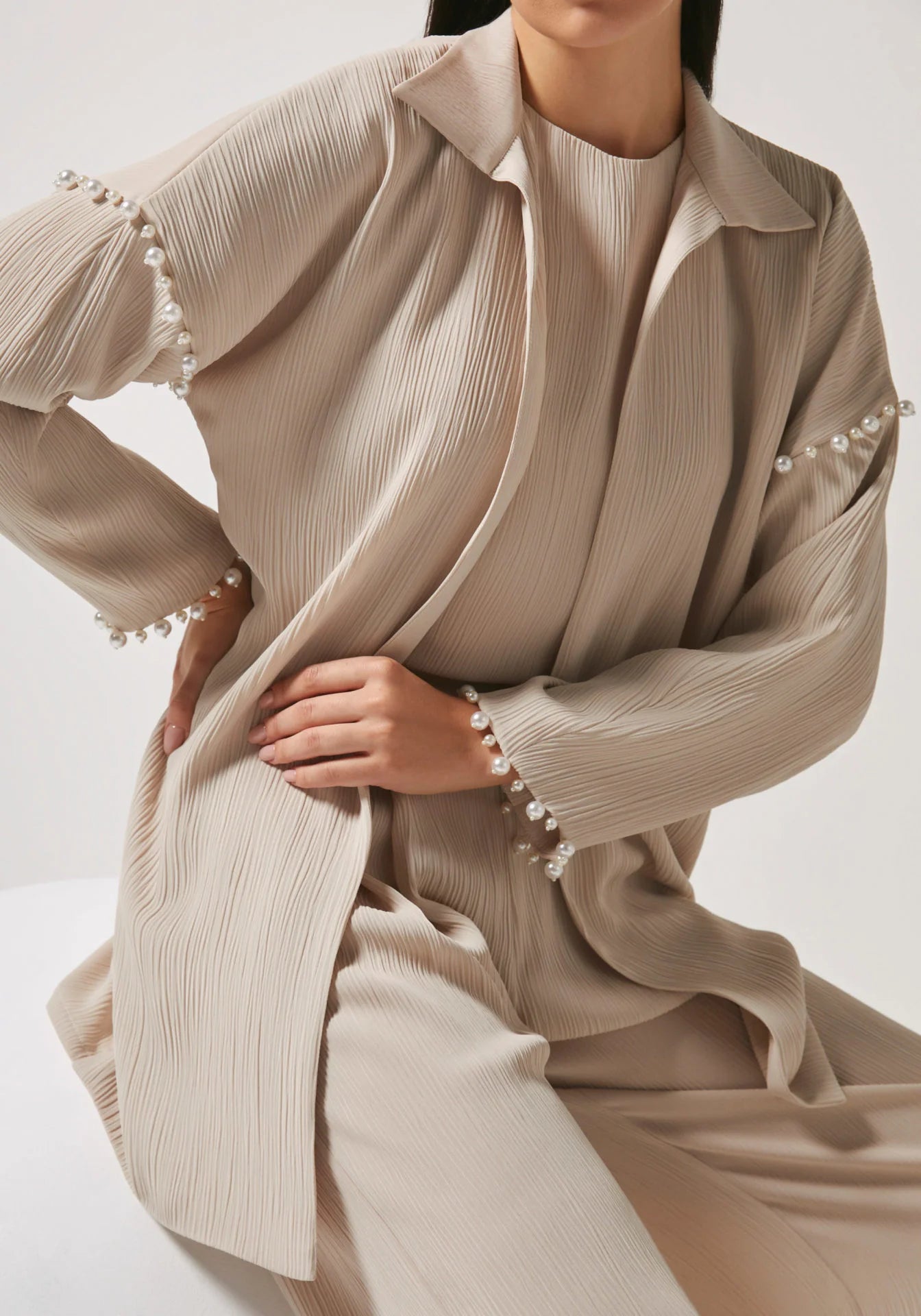 A close-up of a model wearing a beige pleated long-sleeve collared jacket with pearl embellishments along the cuffs, paired with matching pleated trousers.