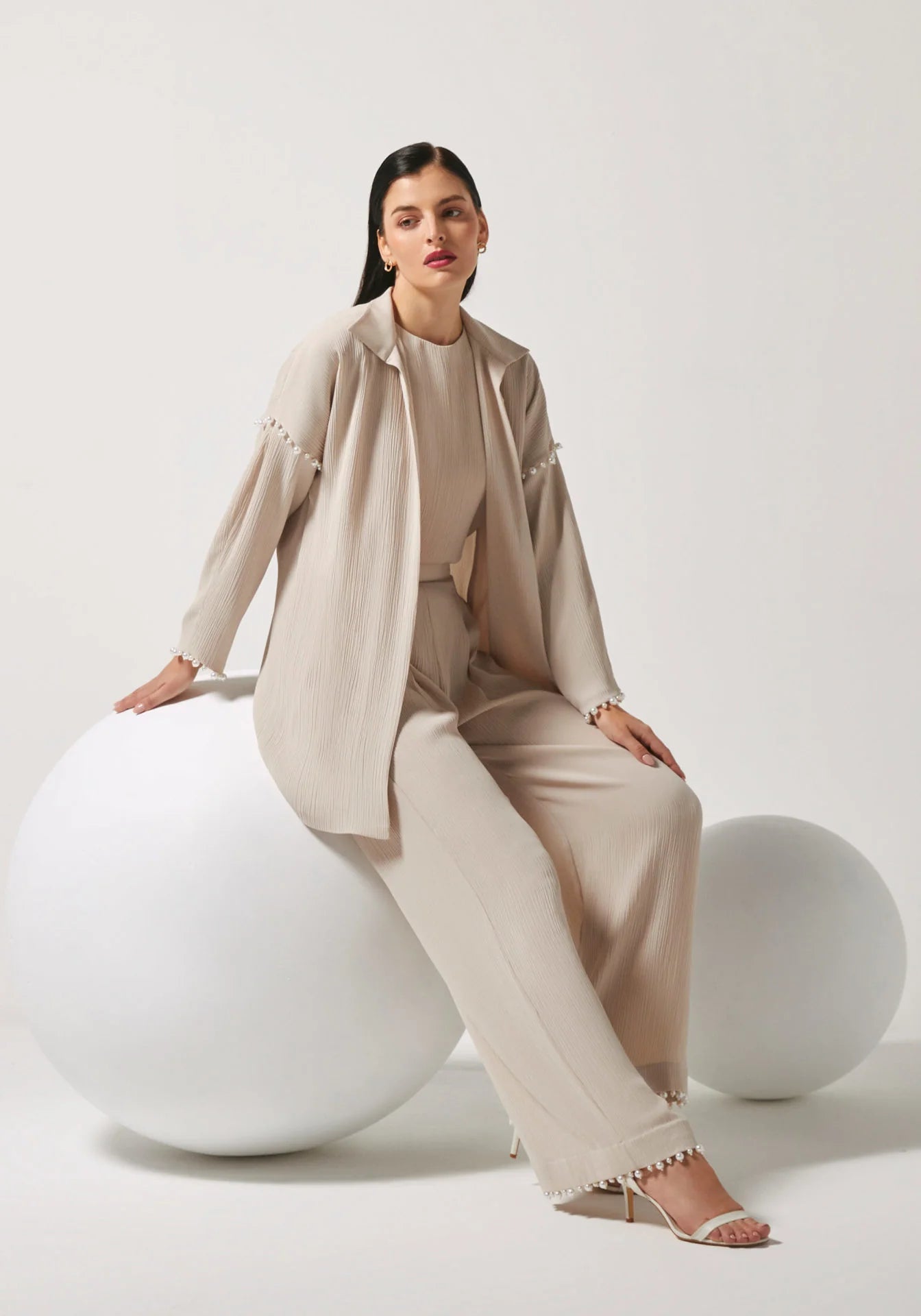 A woman models a beige two-piece linen set consisting of a long-sleeve button-up shirt and matching wide-leg trousers, styled with white heeled sandals and seated on a large white sphere against a light gray studio background.