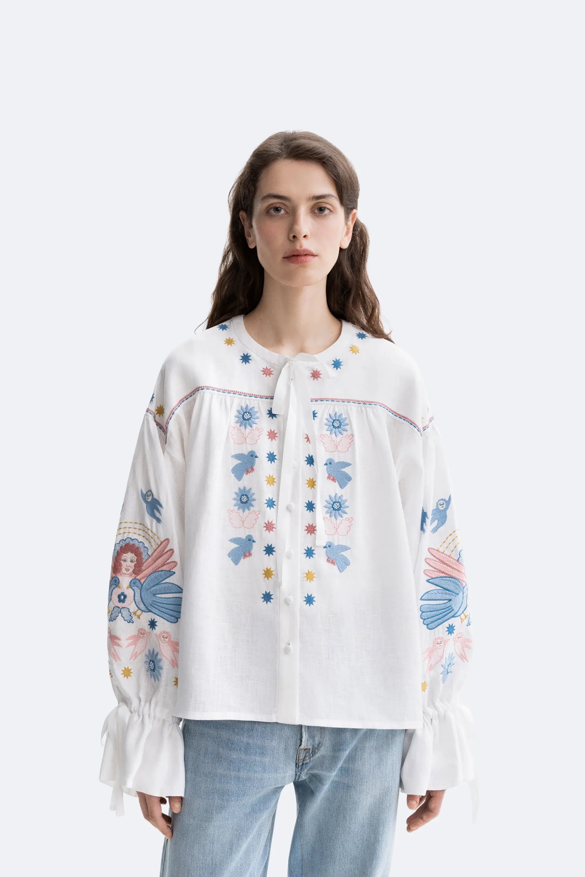 A woman modeling a white long-sleeve blouse with colorful embroidered floral and bird motifs, paired with light blue jeans, against a plain light gray studio background.