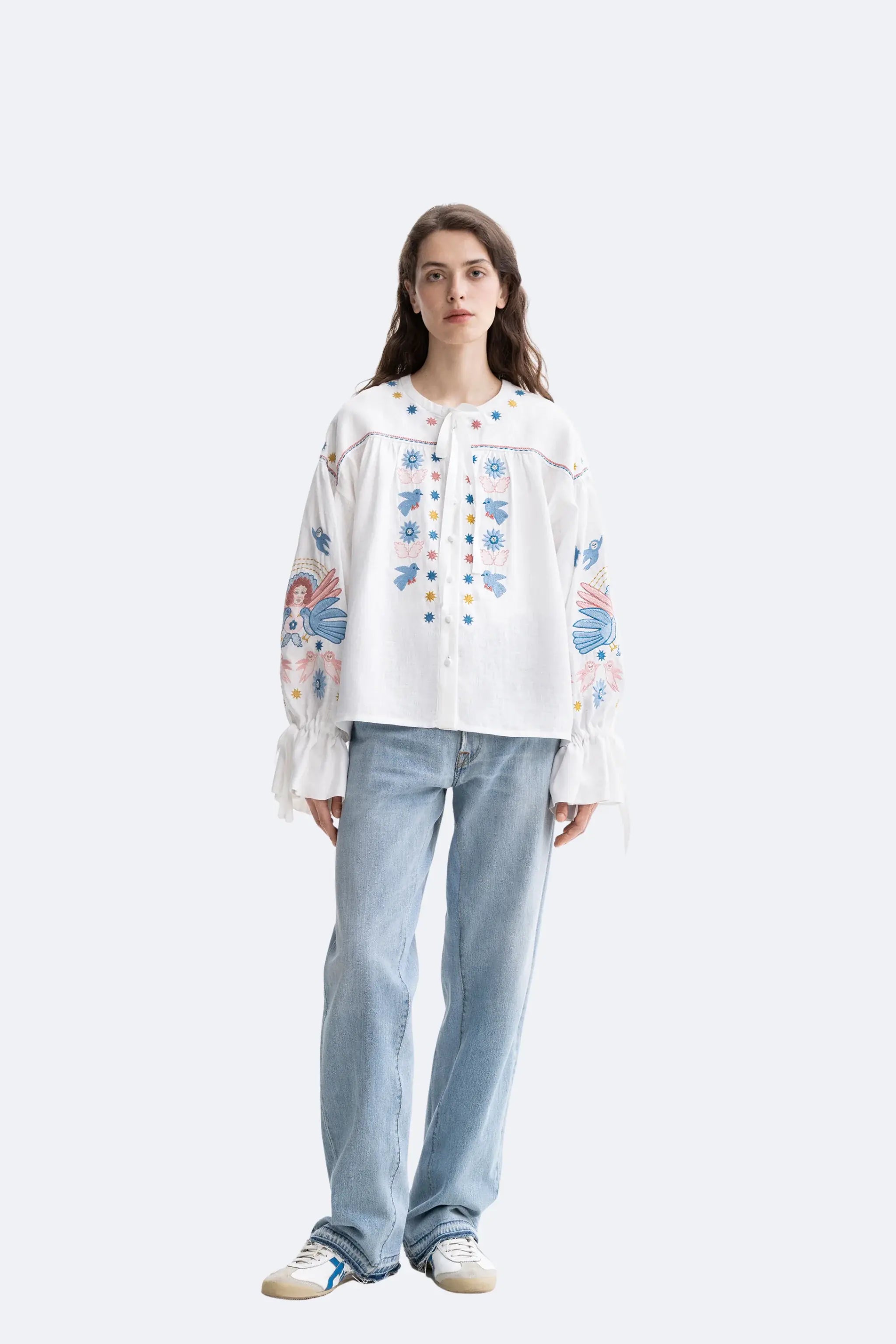 A model stands against a light gray studio background wearing a white embroidered blouse with colorful floral and geometric patterns, light blue straight-leg jeans, and white sneakers with blue accents.