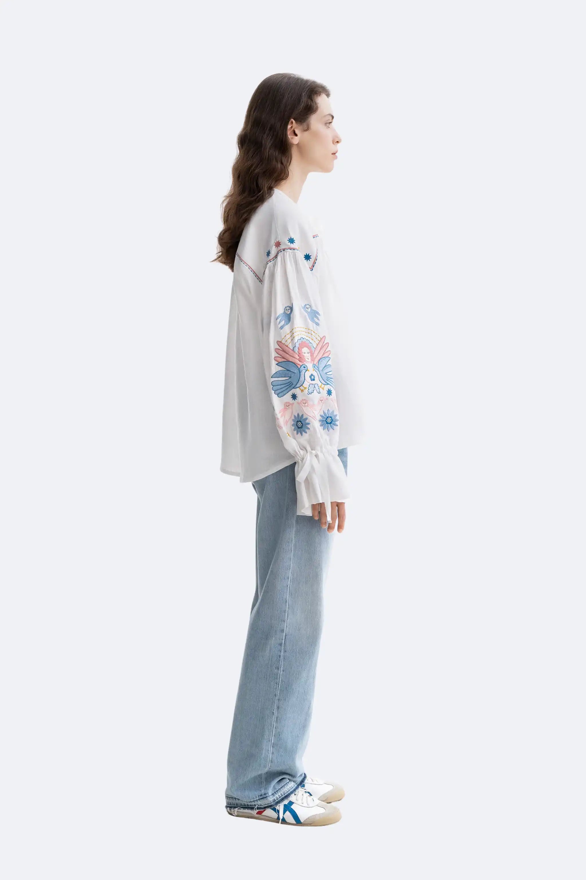 A woman stands in profile wearing a white long-sleeve top with colorful embroidered floral and butterfly motifs on the sleeves, paired with light blue wide-leg jeans and white sneakers, against a plain light gray studio background.