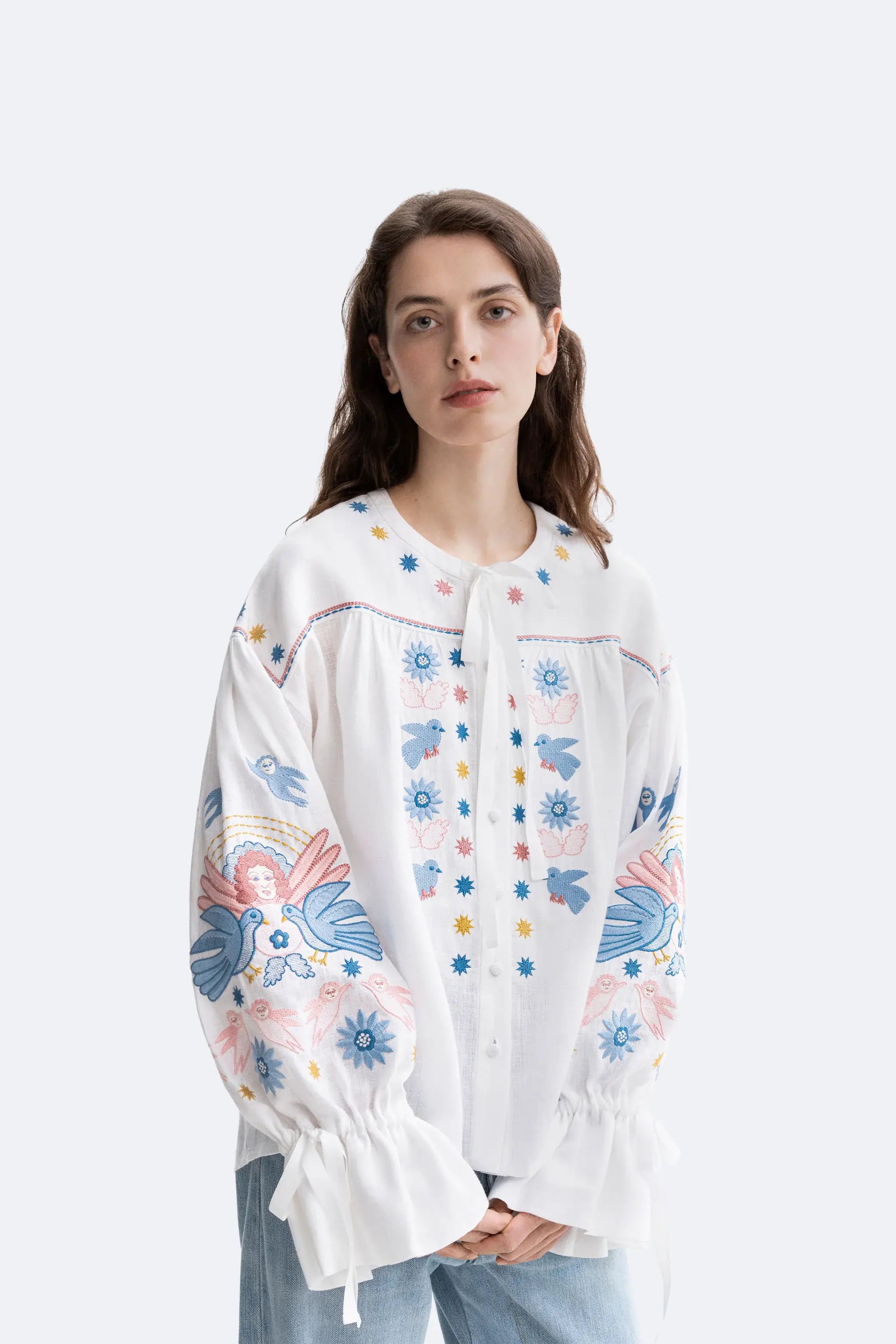 A woman wearing a white embroidered blouse with colorful floral and bird motifs, paired with light blue jeans, against a plain light gray studio background.