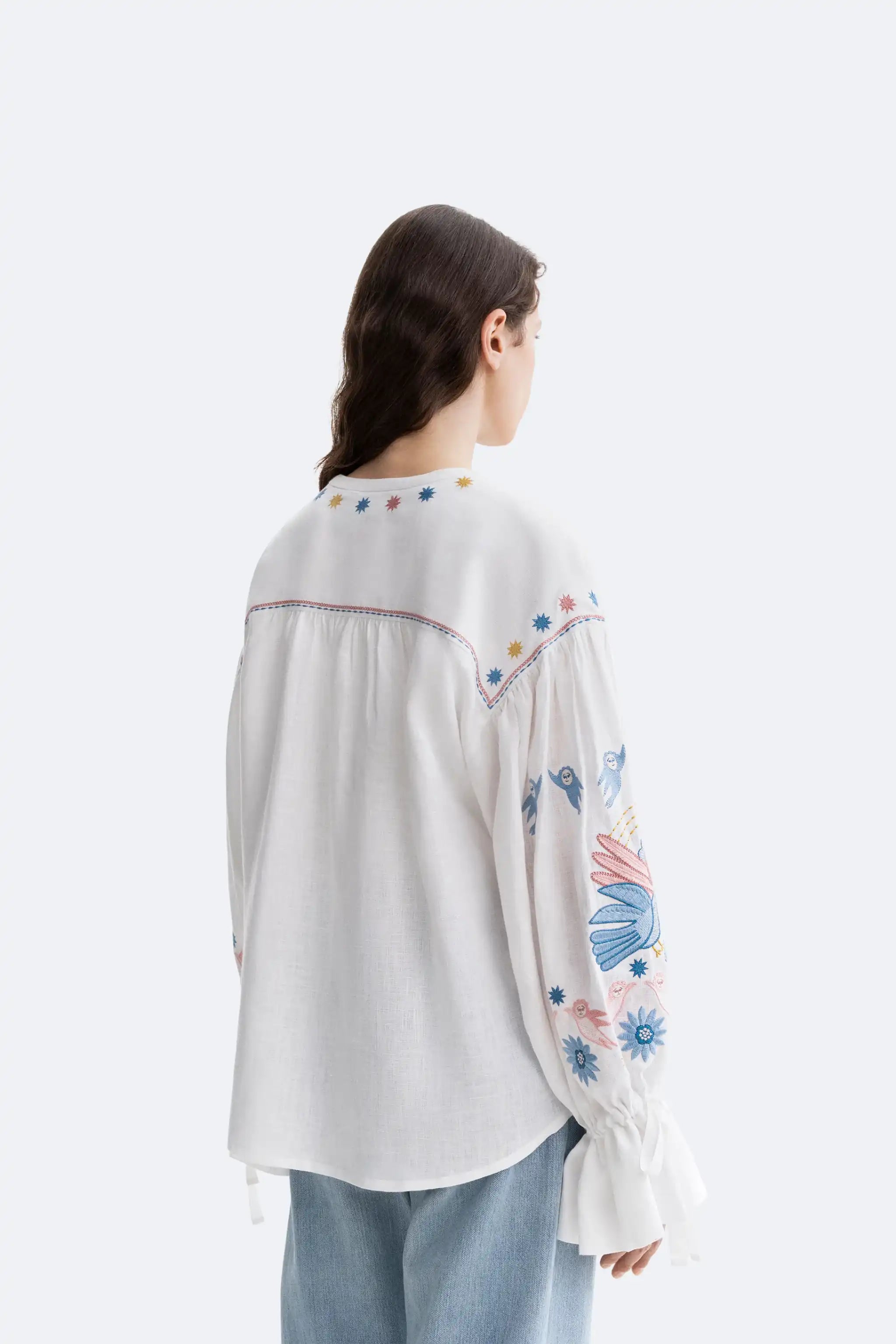 A woman shown from the back wearing a white embroidered blouse with colorful floral and geometric details on the sleeves and neckline, paired with light blue jeans, against a plain light gray studio background.