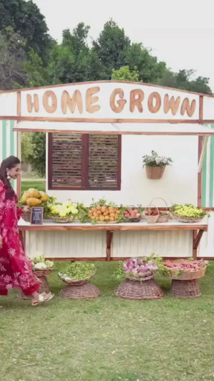 Woman in a red traditional outfit holding an orange in front of a table with fruits and vegetables.