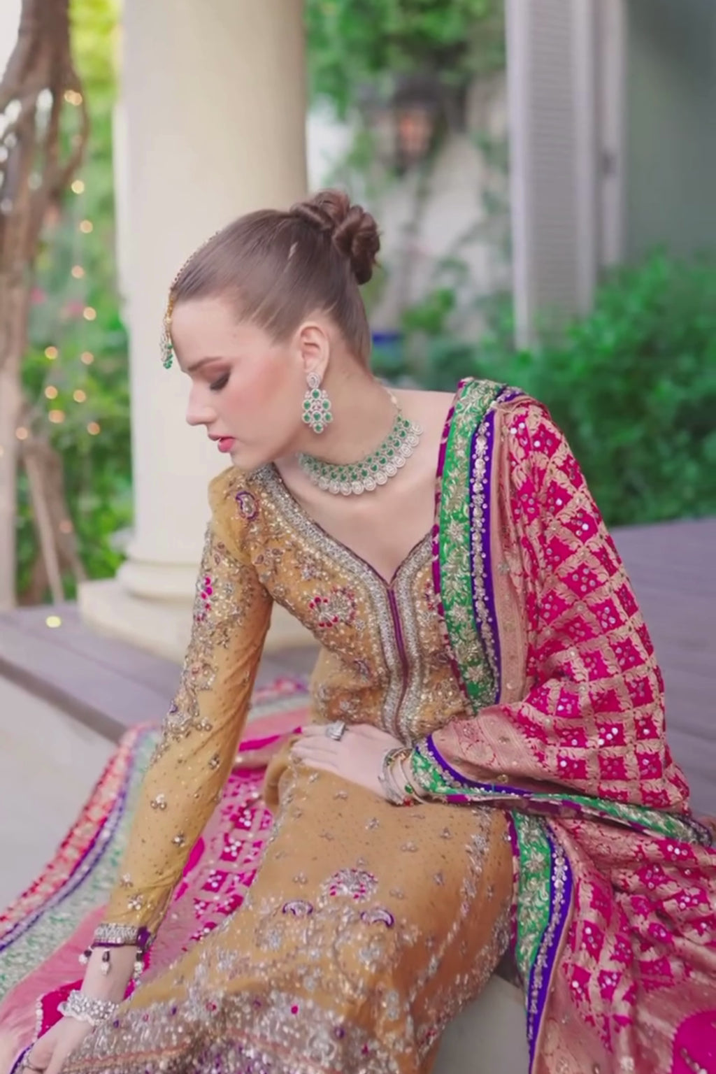 Woman in traditional embroidered banarsi silk gold brown and red color outfit with jewelry, sitting outdoors.