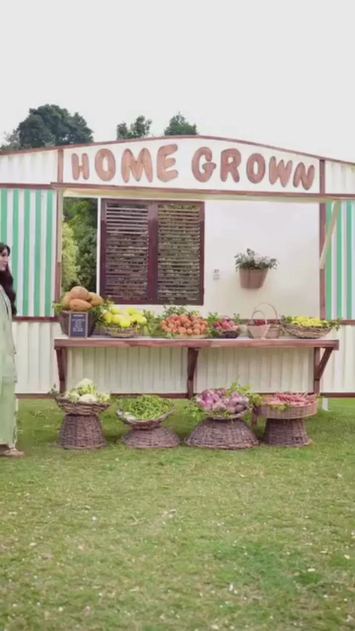 Woman in a light green traditional outfit standing in front of a market stall with fruits and vegetables.