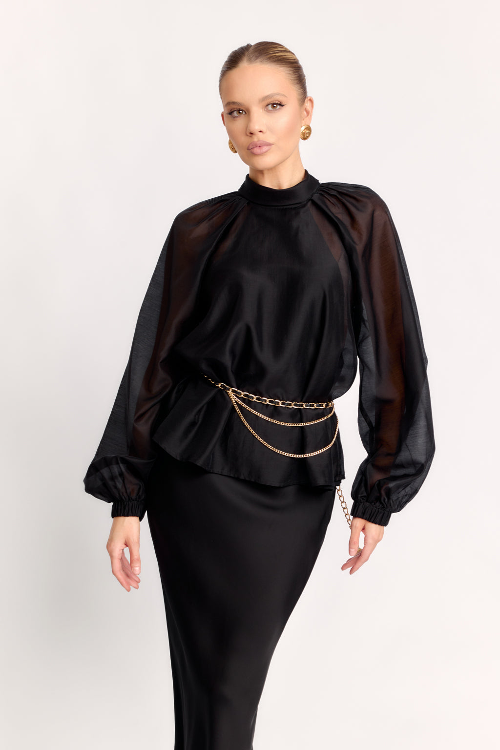 Woman wearing a black blouse with sheer sleeves and a gold chain belt on a white background