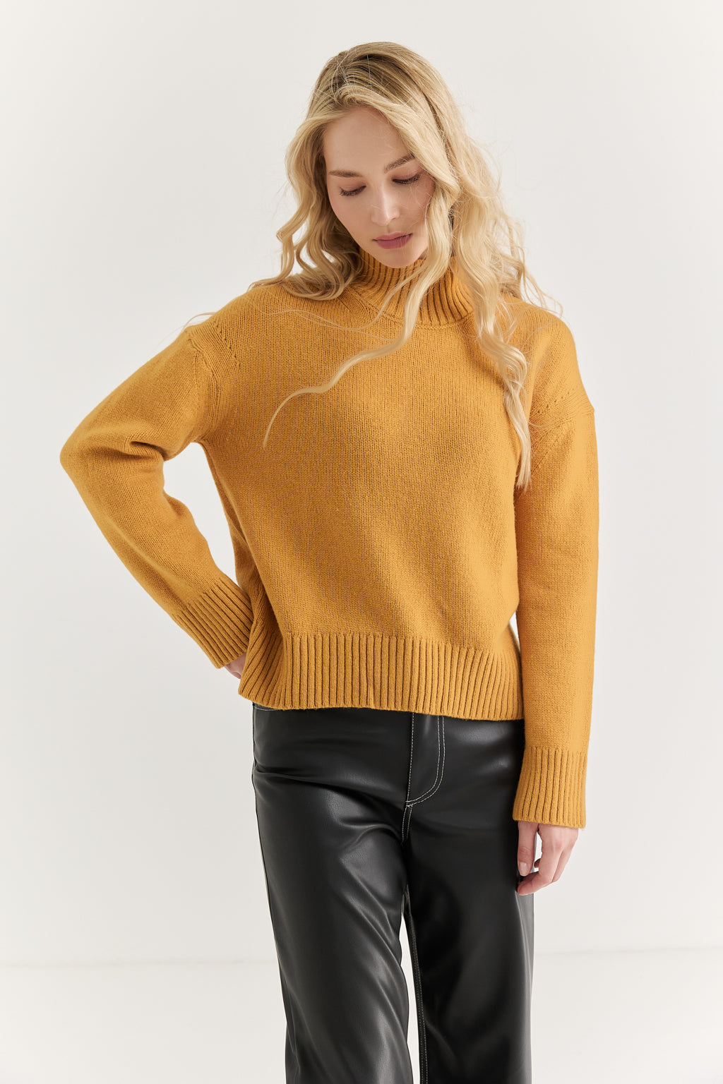 Woman wearing a mustard yellow sweater and black leather pants on a white background
