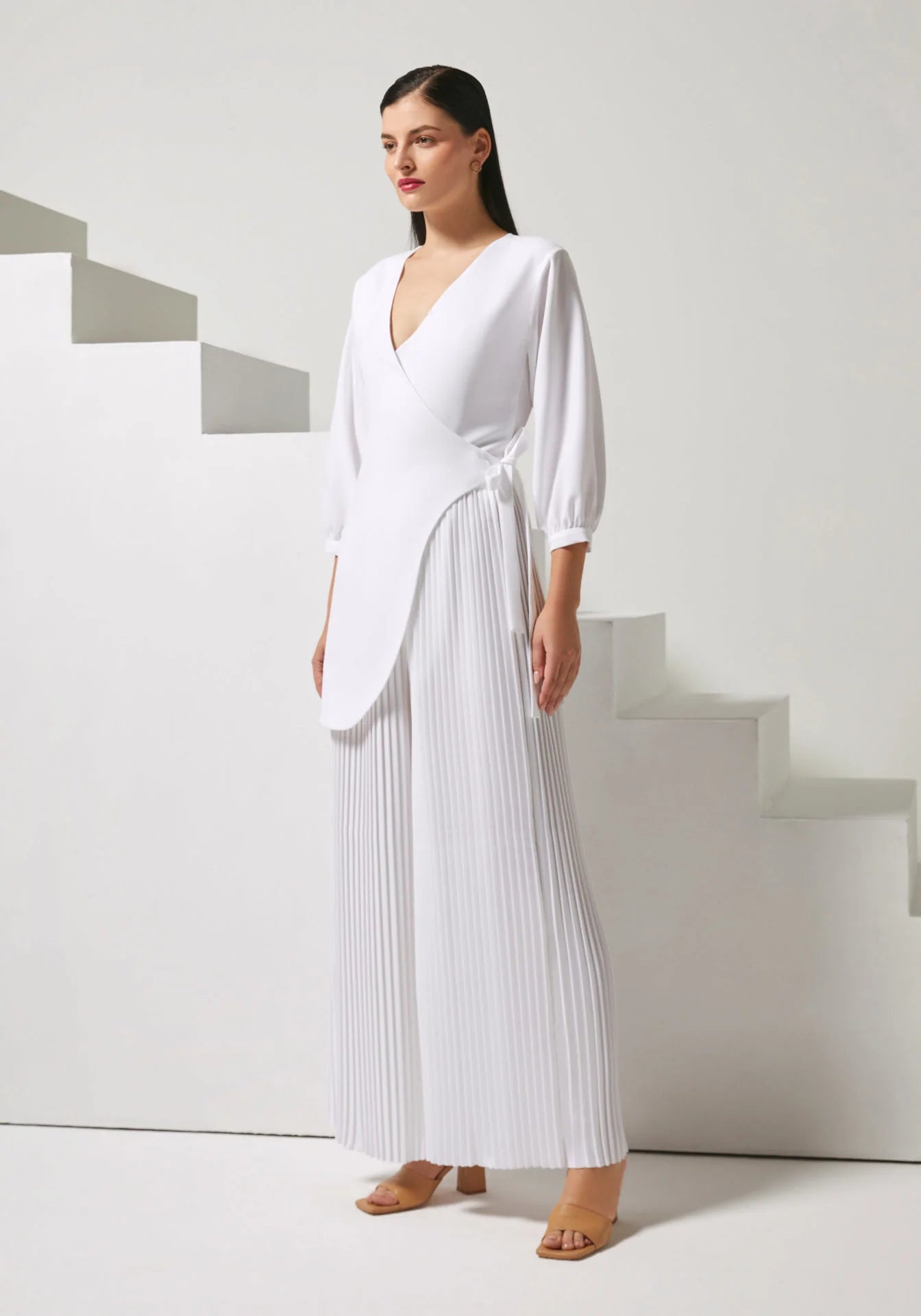 A woman modeling a white wrap-style midi dress with long puff sleeves and a pleated skirt, standing in front of minimalist white steps.