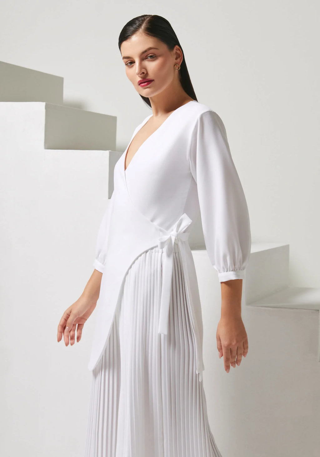 A woman modeling a white wrap-style midi dress with long balloon sleeves, a deep V-neckline, and a pleated skirt, posed against a minimalist white geometric background.