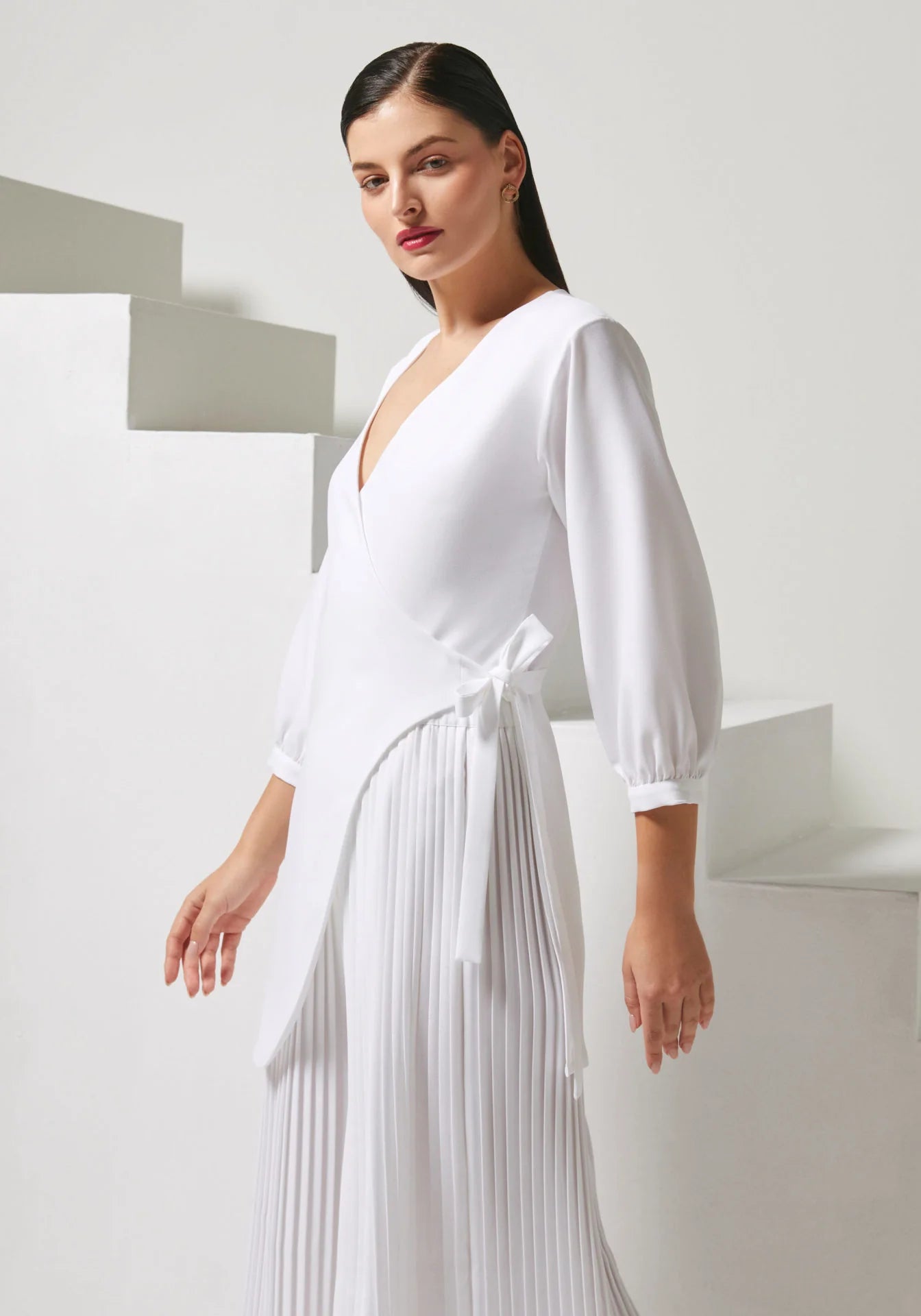 A woman modeling a white wrap-style midi dress with long balloon sleeves, a deep V-neckline, and a pleated skirt, posed against a minimalist white geometric background.