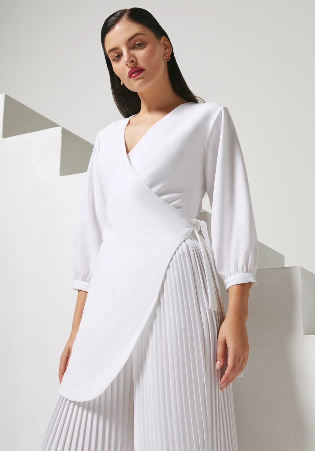 A woman modeling a white wrap-style top with three-quarter sleeves and a pleated white skirt against a minimalist white geometric background.