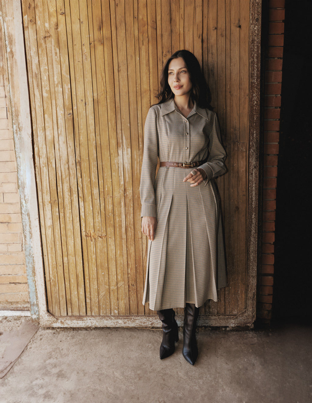 Woman in a long beige coat standing against a wooden wall.
