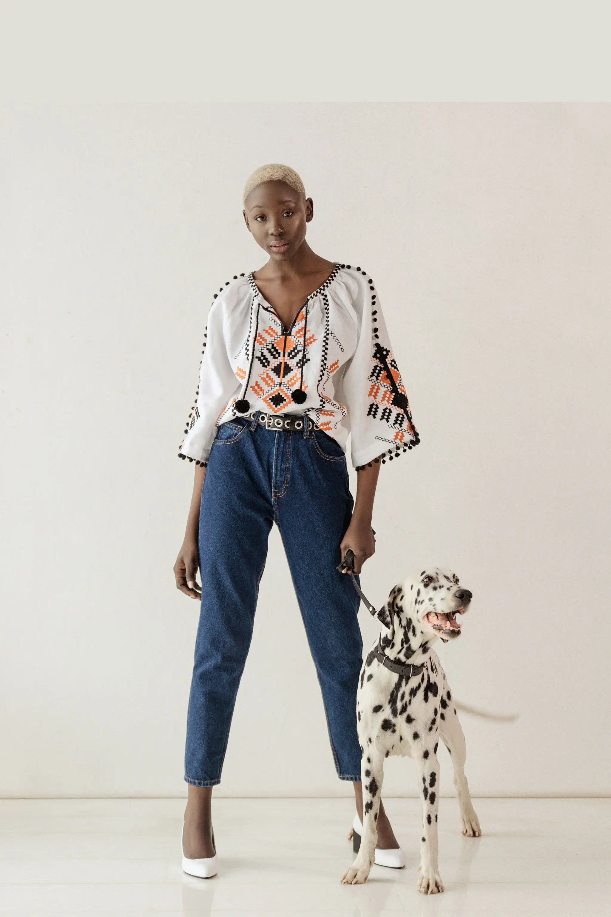 A model stands in a studio wearing a white embroidered blouse with orange and black geometric patterns, blue jeans, and white heels, while holding a leash attached to a Dalmatian dog.