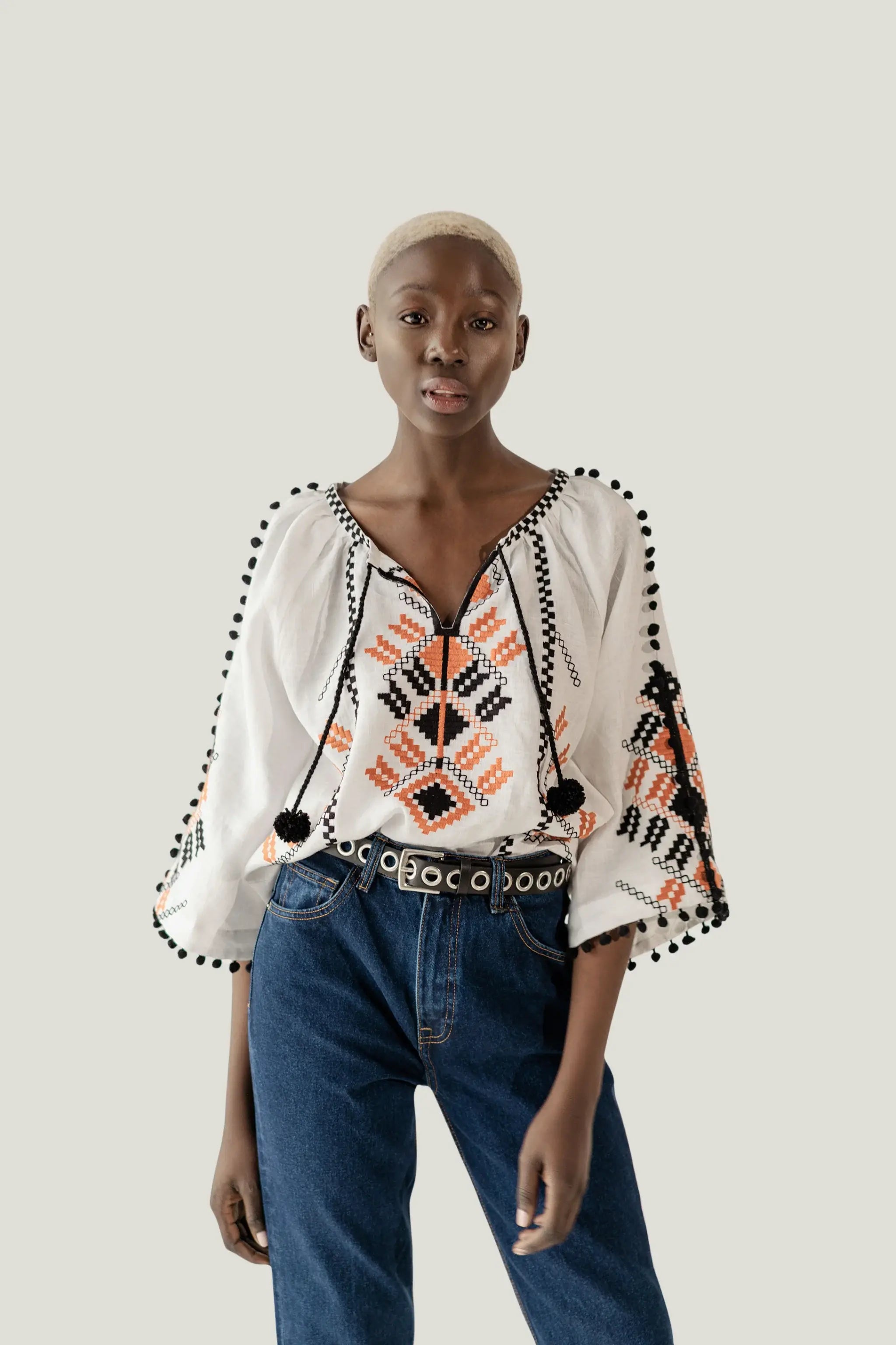 A model wearing a white bohemian-style blouse with black and orange geometric embroidery, pom-pom trim, and drawstrings, paired with blue jeans and a black belt, against a solid light gray background.