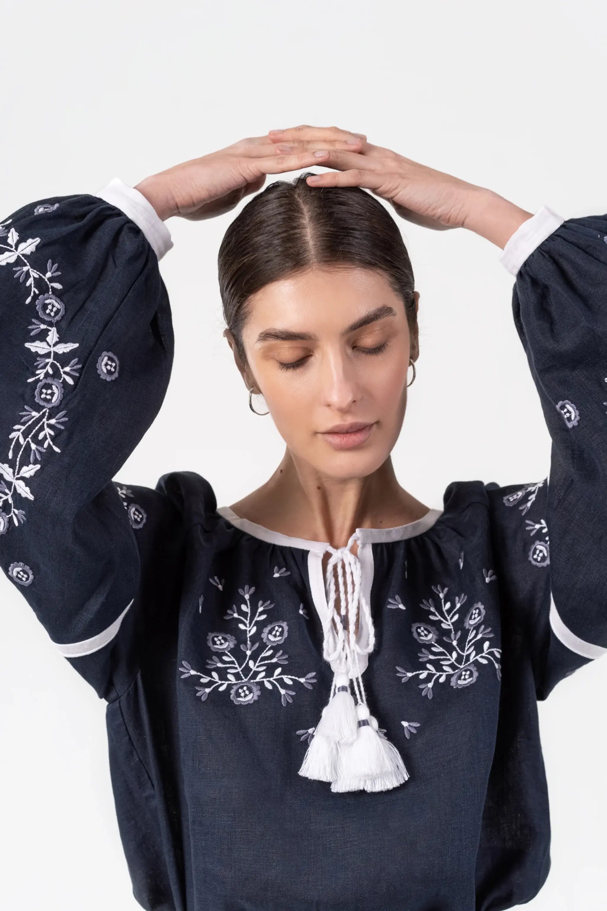 A woman wearing a dark navy blue embroidered blouse with white floral patterns and a white tassel tie at the neckline, posing with her hands on her head against a light gray background.