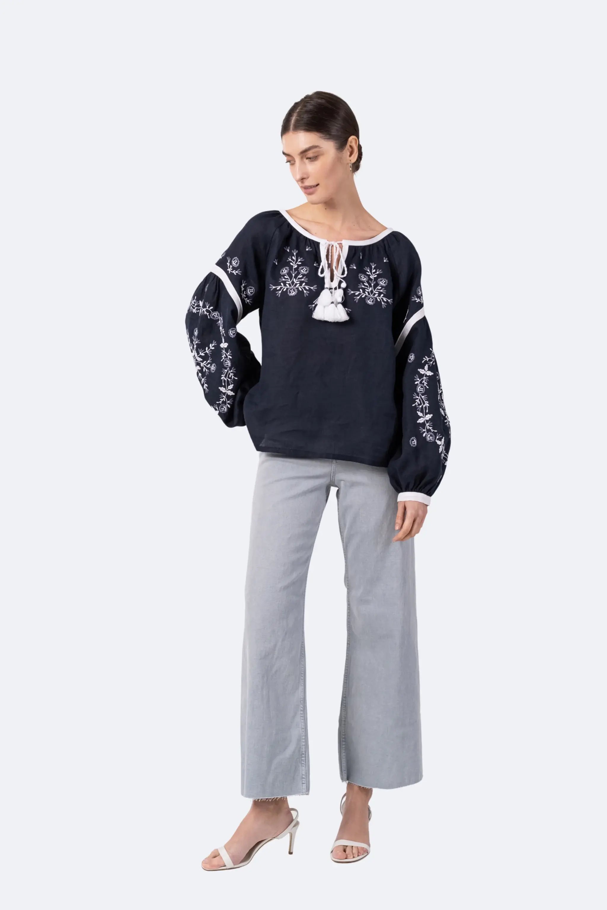 A woman modeling a dark navy blue embroidered blouse with white floral patterns, paired with light gray wide-leg pants and white heeled sandals, against a light gray studio background.