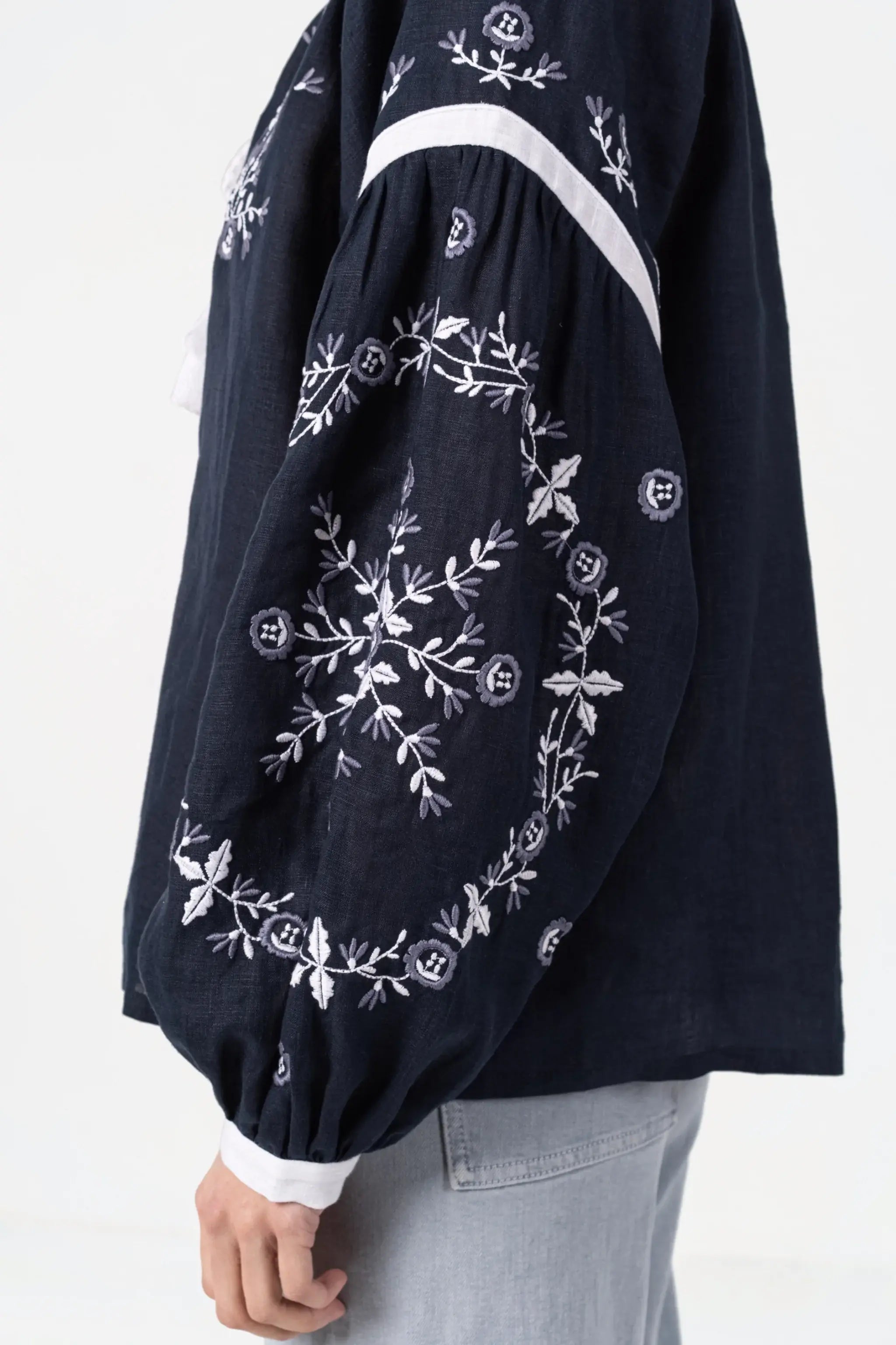 A close-up of a person wearing a dark navy blue long-sleeve blouse with white floral embroidery and a white stripe detail, paired with light gray jeans.