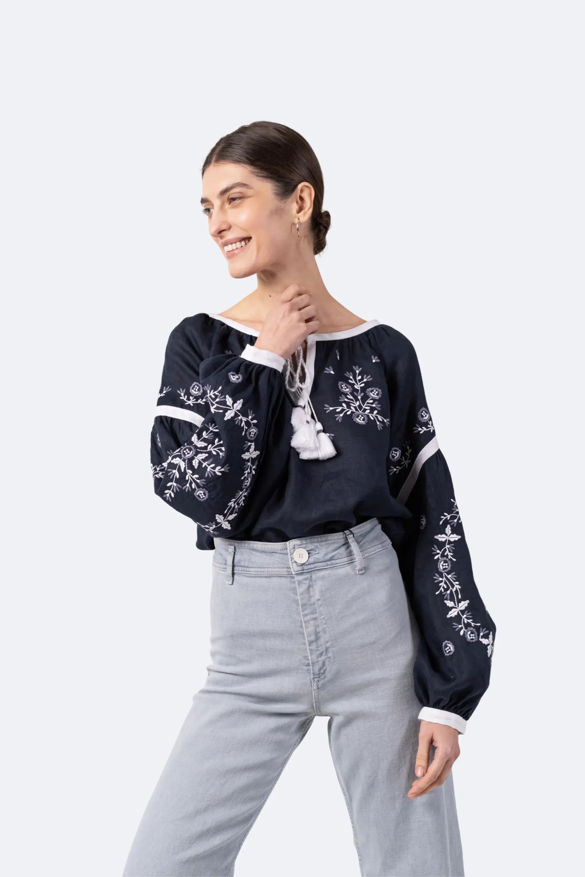 A woman modeling a navy blue off-the-shoulder blouse with white floral embroidery and tie details, paired with light gray jeans, against a solid light gray background.