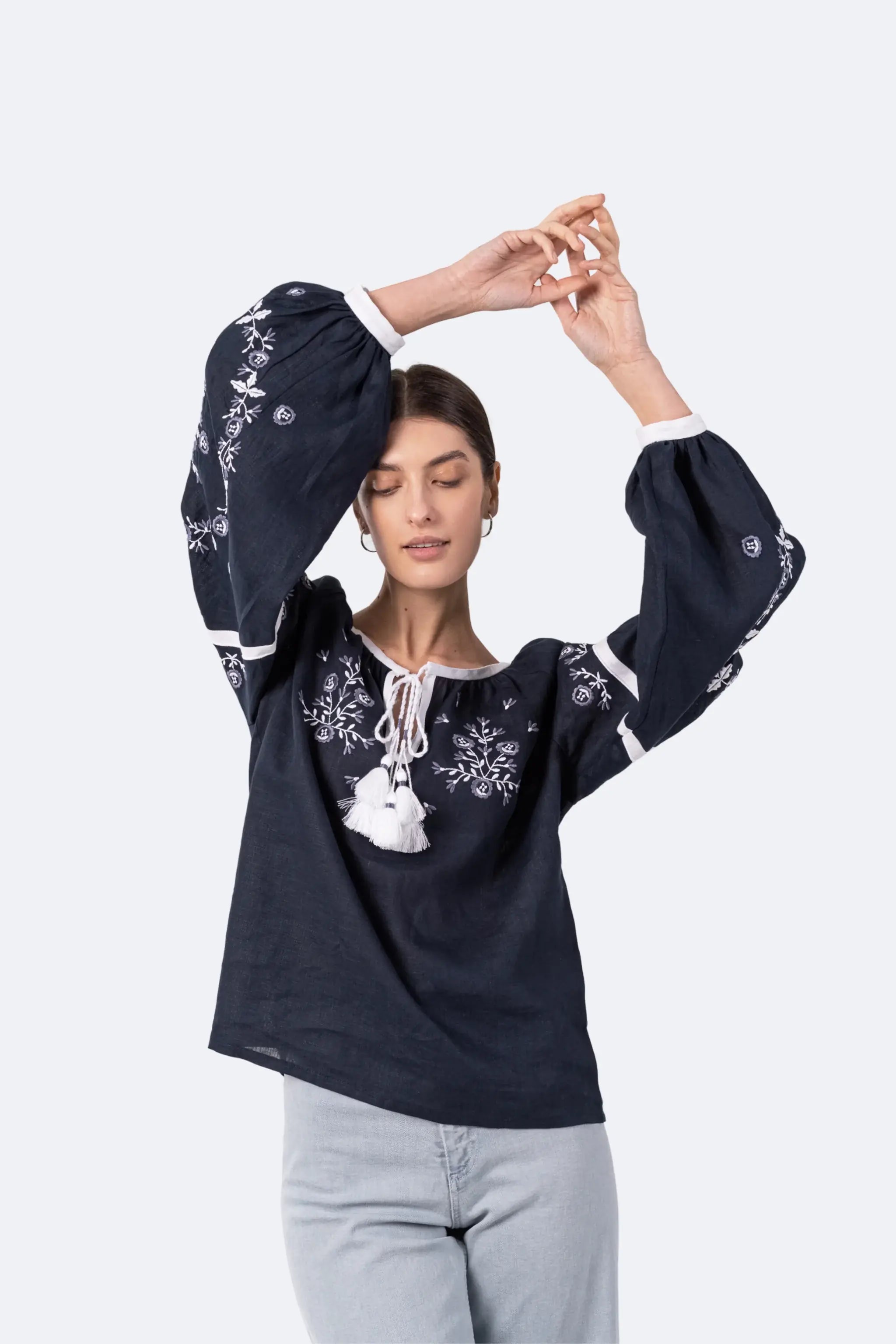 A woman modeling a navy blue long-sleeve blouse with white floral embroidery, a white drawstring tie at the neckline, and white cuffs, paired with light gray jeans against a light gray studio background.