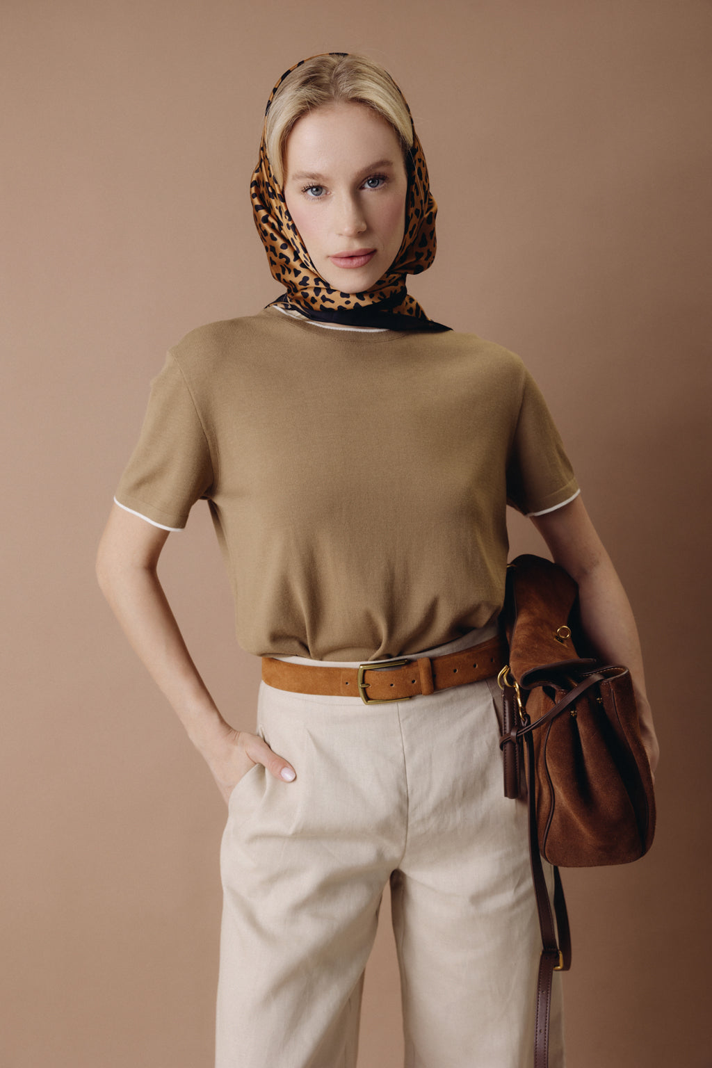 Woman wearing a brown top and beige pants with a leopard print headscarf and brown handbag against a beige background