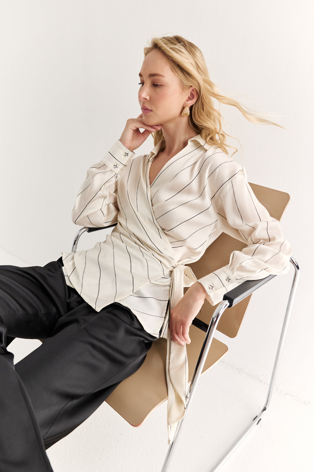 Woman sitting on a chair wearing a cream-colored blouse with black pants against a white background