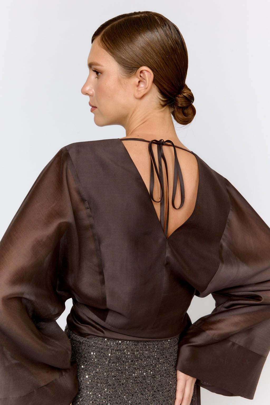 Woman wearing a brown satin blouse with sheer sleeves and a back tie detail on a white background