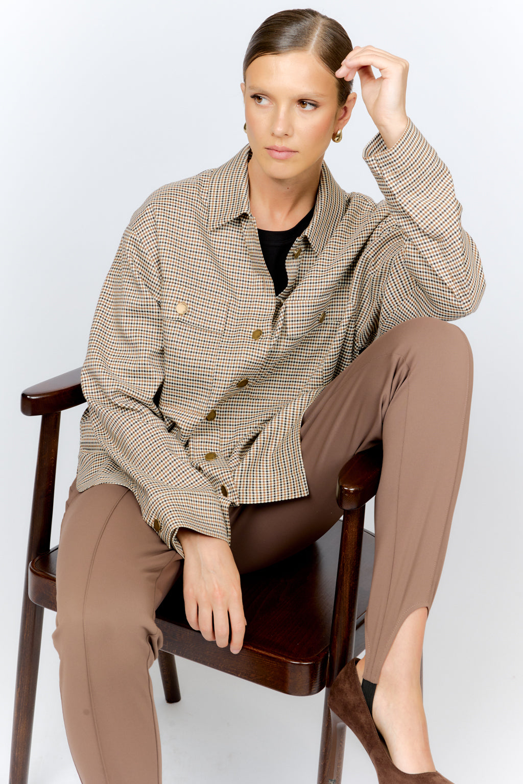 Woman sitting on a chair wearing a checkered jacket and brown pants against a white background