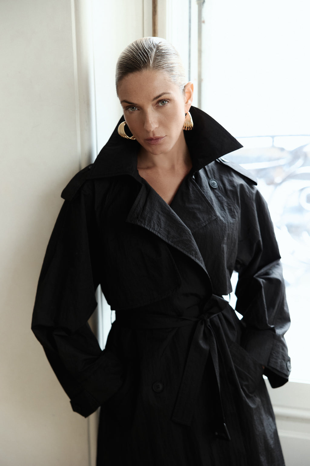 Woman wearing a black coat standing indoors with a window in the background
