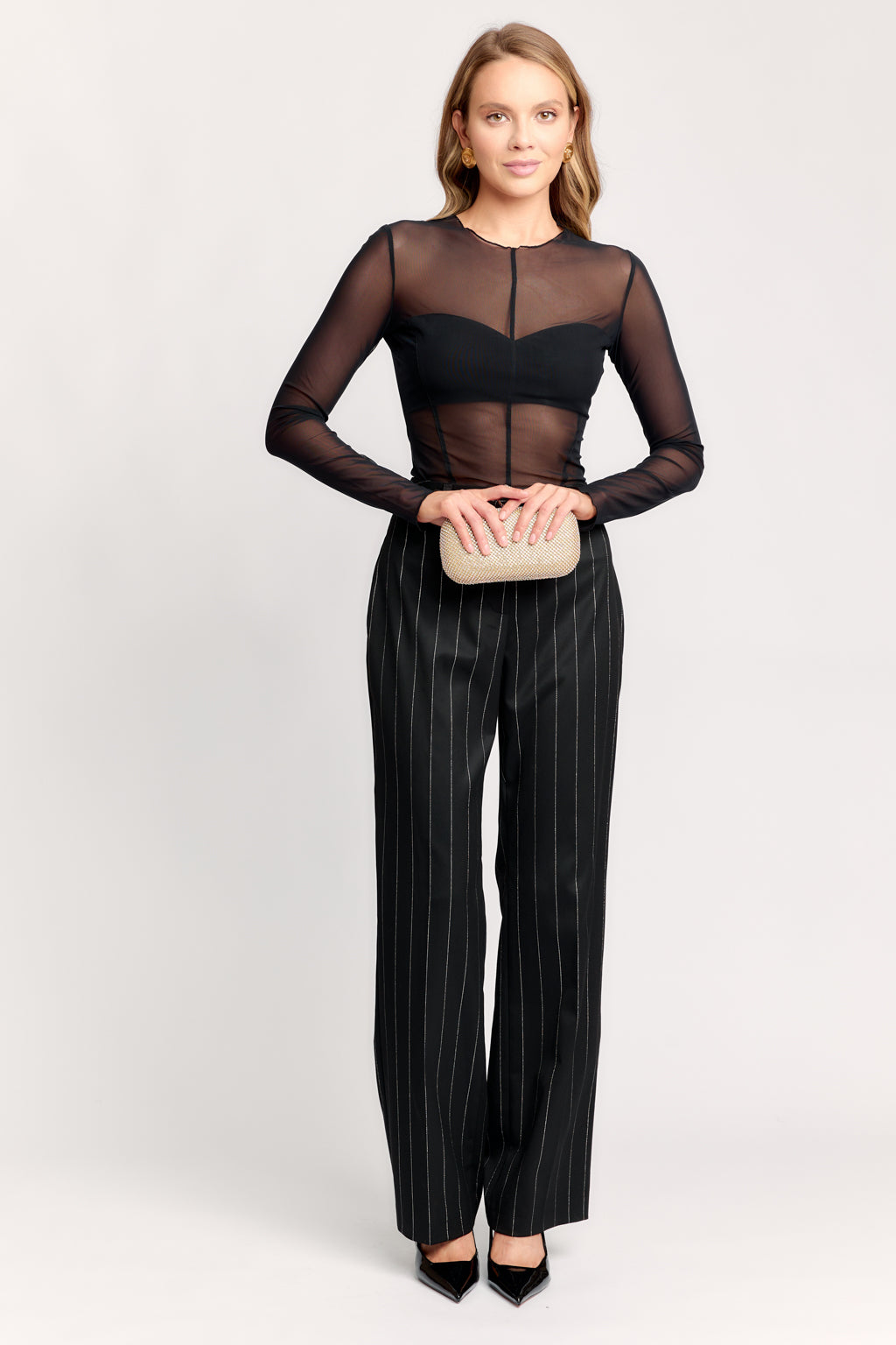 Woman wearing a black sheer top and pinstripe pants holding a beige clutch on a white background