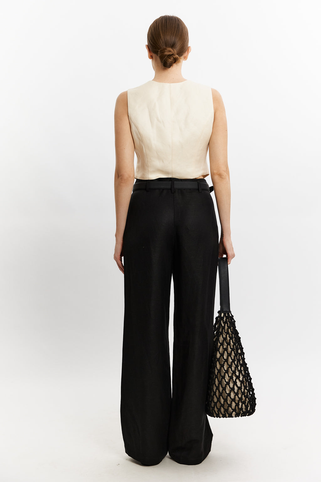 Person wearing a beige sleeveless top and black wide-leg pants, holding a black mesh bag on a white background.