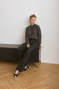 Woman sitting on a radiator wearing a black polka dot blouse and black pants in a minimalistic room.