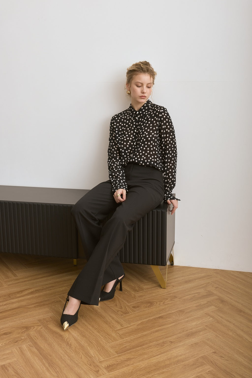 Woman sitting on a radiator wearing a black polka dot blouse and black pants in a minimalistic room.